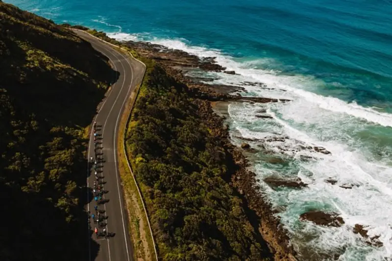 An aerial view of cyclists riding along a winding coastal road that curves alongside a dramatic rocky shoreline. The road features yellow lane markings and cuts through green vegetation-covered terrain. Turquoise ocean water crashes against dark rocky outcrops, creating white foam and waves.