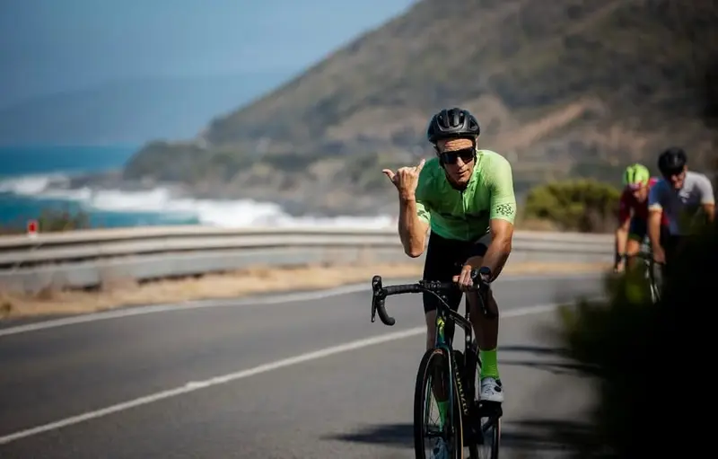 A cyclist in an aerodynamic riding position on a time trial bike gives a thumbs up gesture while riding on a coastal road. The rider wears a bright green jersey, black helmet, and sunglasses. In the background, rolling hills slope down to the ocean, and other cyclists can be seen further back on the road.
